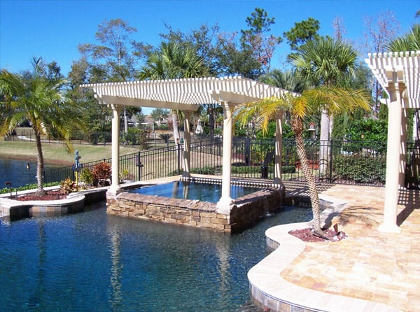 A white pergola over a hot tub with a stone border that is part of a larger pool with a stone deck to one side and a lake in the background on the other side. 